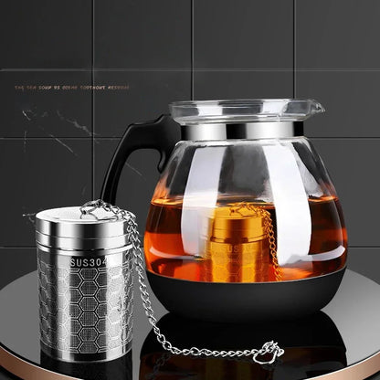 Glass teapot with black handle and metal tea infuser on a dark surface.